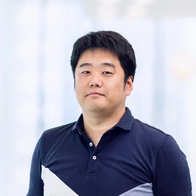 紺色のポロシャツを着たアジア人男性の上半身のポートレートです。男性はカメラをまっすぐ見ており、背景は明るくぼかされています。ビジネスシーンやプロフィール写真に適した落ち着いた雰囲気の画像です。