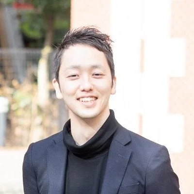 黒いタートルネックと紺色のジャケットを着た若いアジア人男性が、笑顔でカメラを見ているポートレート写真です