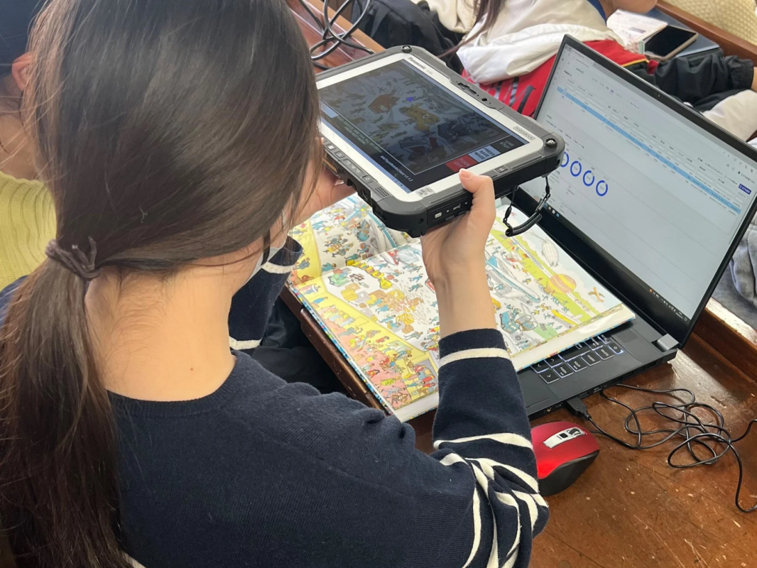 一人の人物が、頑丈なタブレット（Panasonic Toughbook）を『ウォーリーを探せ』のような絵本にかざしている様子を後ろから捉えた画像です。タブレットの画面には絵本の内容が映し出されており、隣にはデータが表示されたノートパソコンも開かれており、デジタルとアナログを組み合わせた学習や研究の場面を示唆しています。