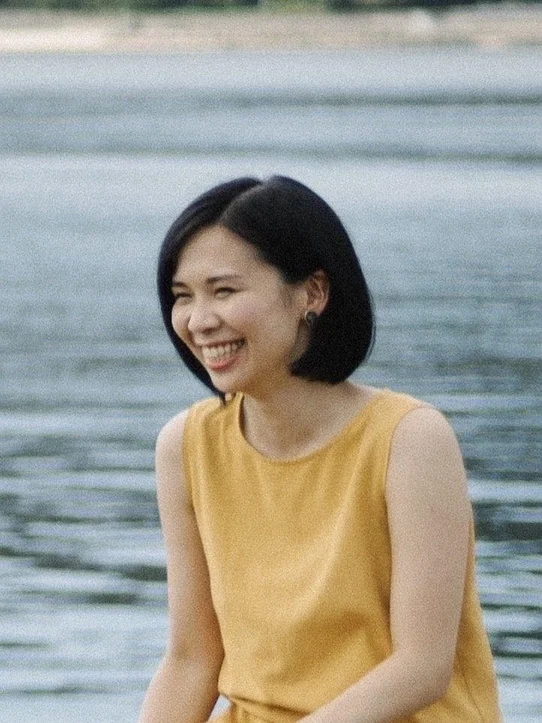 水辺で黄色いノースリーブを着たショートヘアの若い女性が、明るい笑顔を見せているポートレート写真です。背景には穏やかな水面が広がっています。