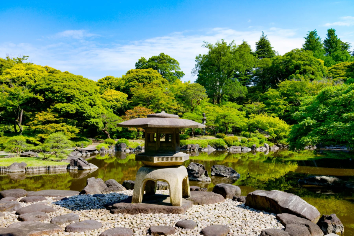 日本の伝統的な庭園の風景。中央に石灯籠が立ち、その奥には緑豊かな木々が映り込む池が広がる