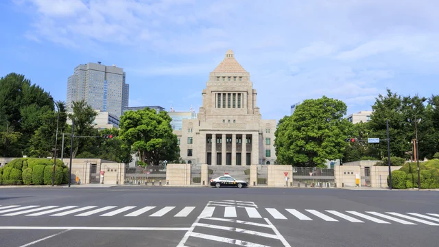 日本の国会議事堂を正面から捉えた日中の風景。手前には横断歩道のある道路があり、中央には警察車両が停車しています。青空のもと、周囲の緑も鮮やかで、日本の政治の中心地を示す一枚です。