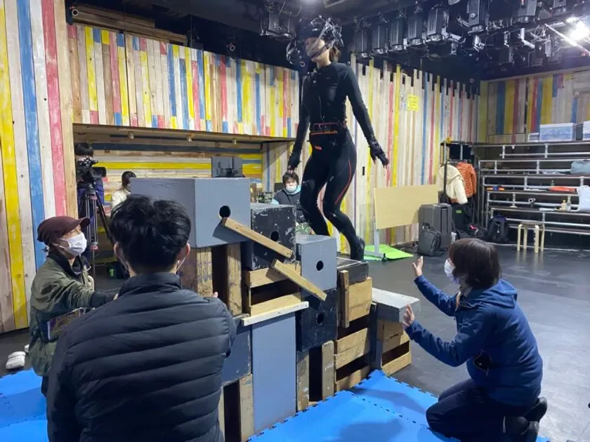 VRヘッドセットとモーションキャプチャスーツを着用した女性が、スタジオ内で積み上げられた台の上でポーズを取っています。周囲には撮影スタッフらしき人々がおり、撮影現場の様子が伺えます。