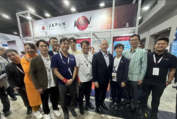 展示会会場の「JAPAN PAVILION」ブース前で、男女混合のグループが集合写真を撮影している。ビジネスイベントの参加者や関係者と思われる人々が笑顔で並んでいる。