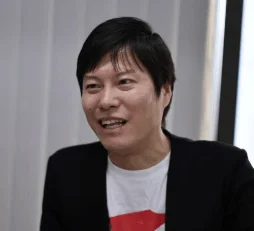 黒いジャケットと白いTシャツを着たアジア系の男性が、笑顔で横を向いているポートレート写真です。会話中かインタビュー中のような雰囲気で、親しみやすい表情をしています。