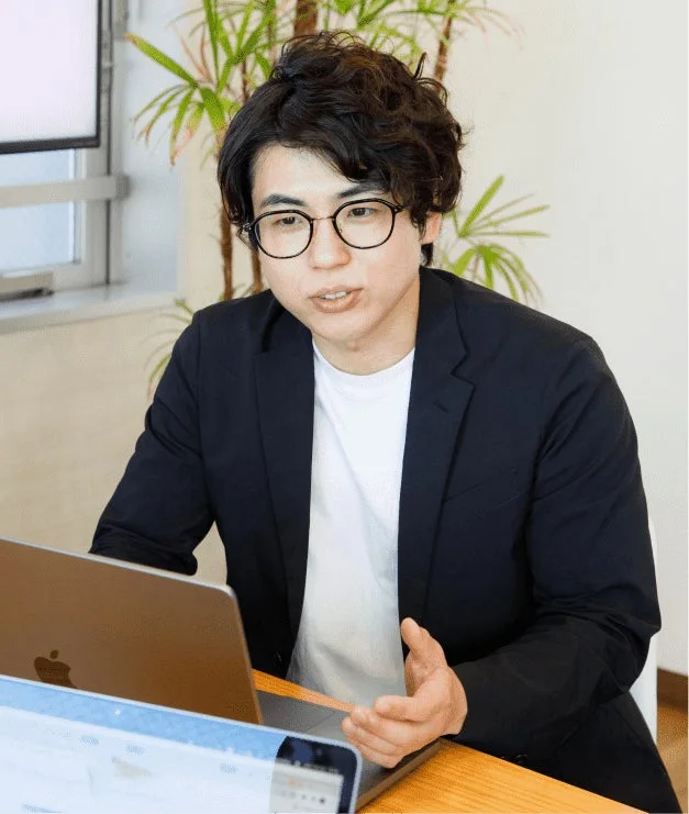 メガネをかけた若い男性が、オフィスらしき場所でノートパソコンを前に座り、何かを説明しているかのように話している様子を捉えた画像です。背景には観葉植物が見えます。