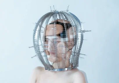 顔に複雑な金属製のケージ状の頭飾りを装着した人物のポートレートです。頭飾りにはスパイクが付いており、顔が複数に見えるような視覚効果があります。