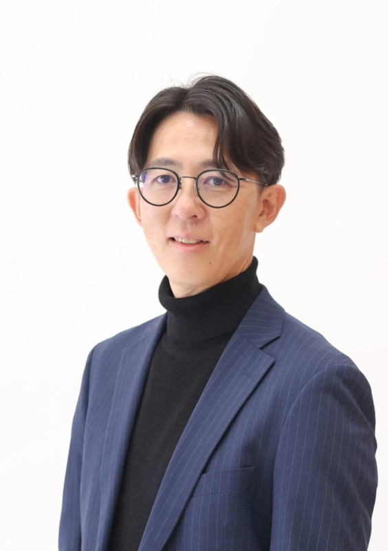 青いストライプのスーツジャケットと黒いタートルネックを着用し、メガネをかけた男性の上半身ポートレート。