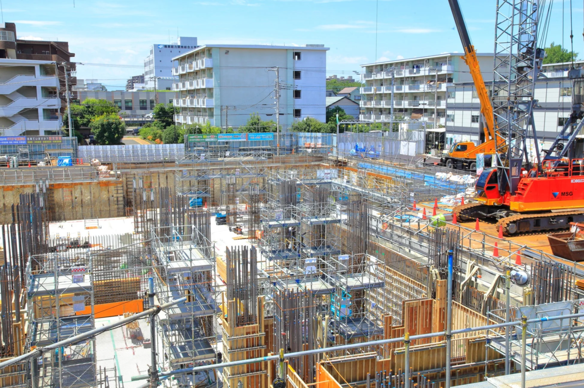 晴れた日に広がる大規模な建設現場の様子。深く掘られた基礎部分には鉄筋や足場が組まれ、赤い大型クレーンが稼働している。周囲にはマンションや大学の建物が見える都市の風景。