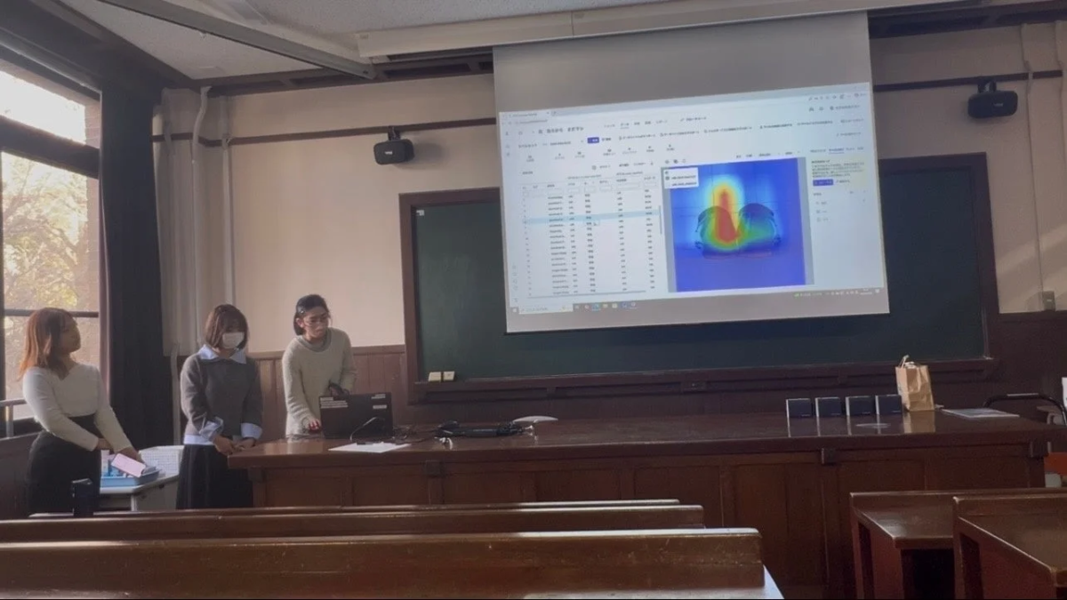 教室で3人の女性がプレゼンテーションを行っています。プロジェクターにはデータテーブルとヒートマップが表示されており、学術的な発表やセミナーの様子を捉えています。発表者はノートパソコンを操作しています。