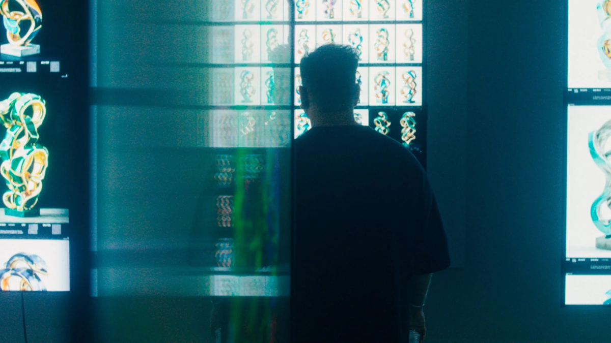 暗い空間で、一人の人物が多数のスクリーンに表示された抽象的なデジタルアート作品を鑑賞している。緑や青の複雑な形状が特徴で、現代的なアート展示やクリエイティブな環境を示唆している。