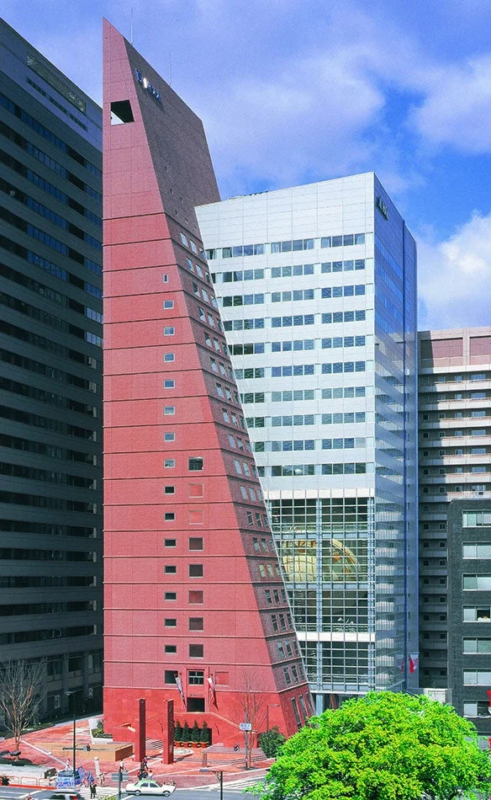 赤い楔形をした特徴的な高層ビルが、他の近代的なビル群に囲まれてそびえ立つ都市の景観です。