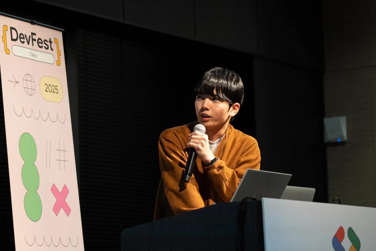 「DevFest Tokyo 2025」のイベントで、若い男性がマイクを持って壇上でプレゼンテーションを行っている。