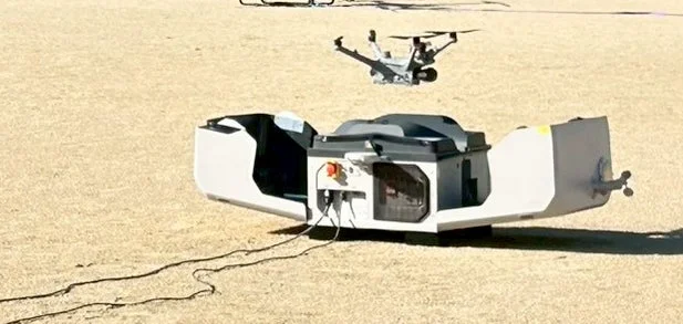 ドローンが地上ステーションの上でホバリングしている様子を捉えた画像です。地上ステーションは充電や自動離着陸のための設備と考えられ、砂地のような場所で運用されていることが伺えます。