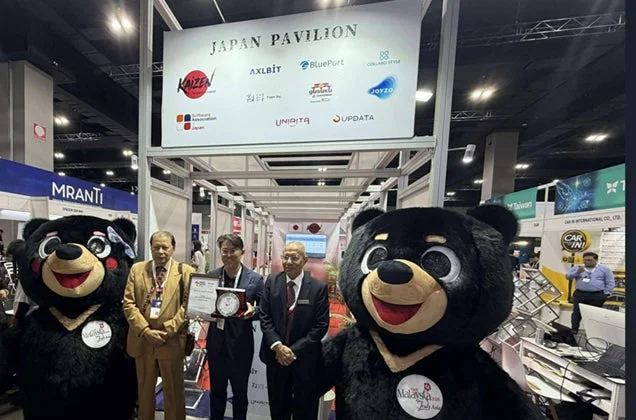 展示会のジャパンパビリオンブースにて、日本のビジネス関係者3名がマレーシアのクマのマスコット2体と共に立ち、表彰状を受け取っている様子。看板には複数の日本企業ロゴが見える。