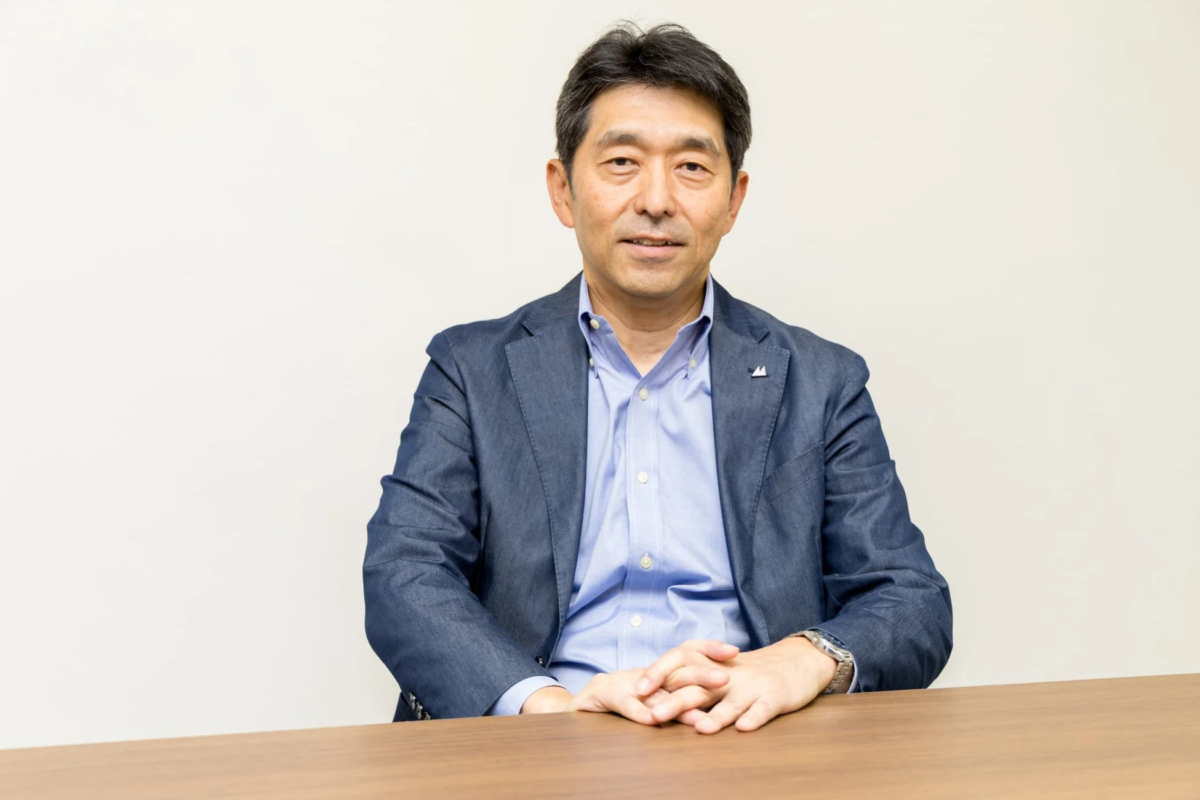 白い背景の前に座り、紺色のジャケットと水色のシャツを着た中年のアジア人男性が、カメラに向かって優しく微笑んでいます。両手を組んで木製のテーブルに置いており、落ち着いた雰囲気のポートレート写真です。