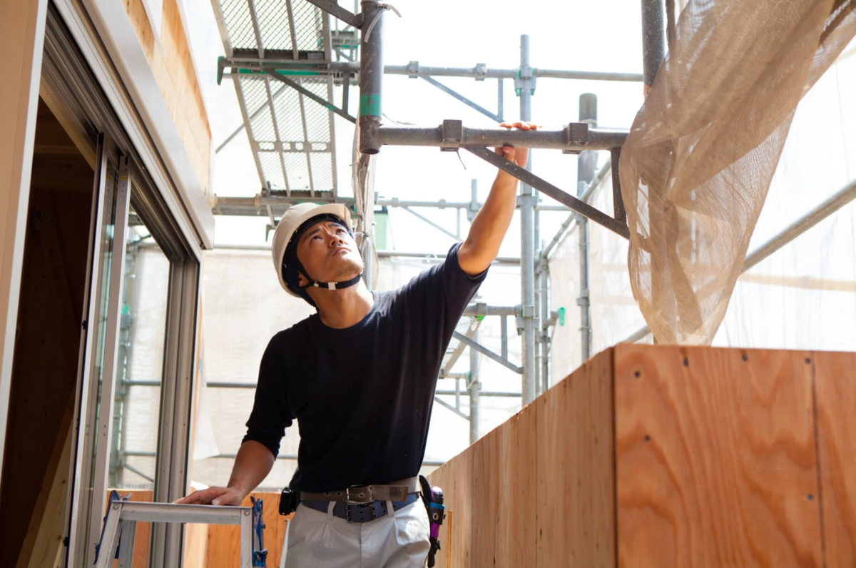 ヘルメットを着用した男性作業員が建設現場の足場を見上げている様子です。真剣な表情で作業に取り組む姿が捉えられています。