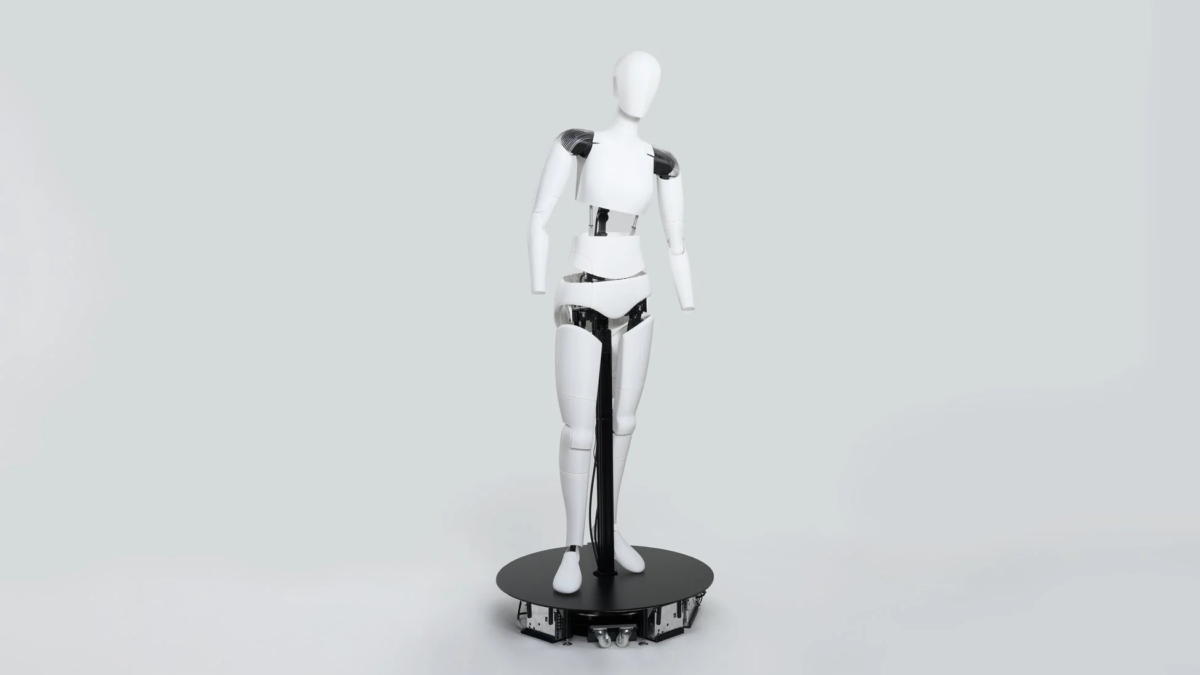 白いヒューマノイド型ロボットが、関節部分に黒いメカニズムを露出させながら、シンプルな灰色の背景に立っています。下半身は車輪付きの黒い円形台座に固定されており、現代的で未来的なデザインが特徴です。