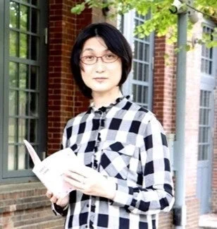 眼鏡をかけた女性が、黒と白のチェック柄シャツを着て本を手に持ち、レンガ造りの建物の前で立っているポートレート写真です。