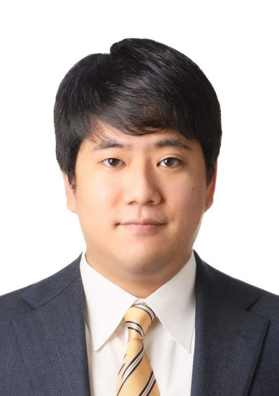 白い背景の前で、ダークスーツとストライプのネクタイを着用したアジア人男性のバストアップポートレートです。真顔で正面を向いています。