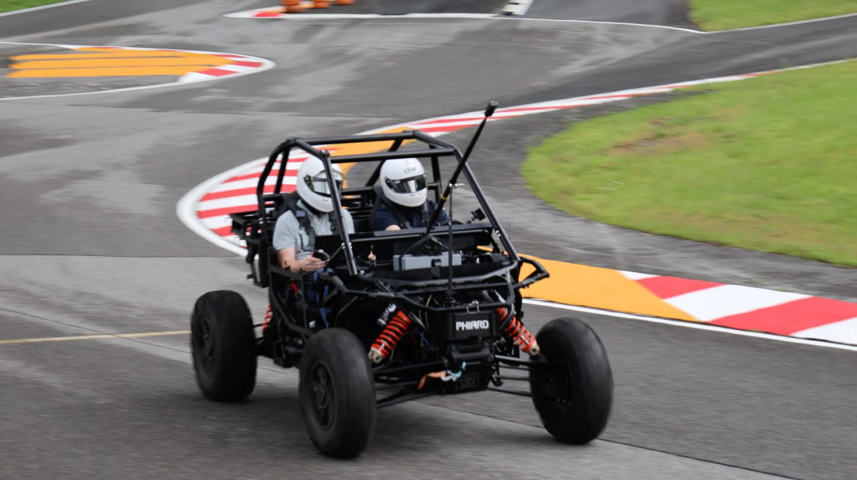 ヘルメットを着用した2人が黒いバギーのような乗り物に乗って、カーブのあるサーキットを走行している様子です。スピード感があり、モータースポーツを楽しんでいるようです。