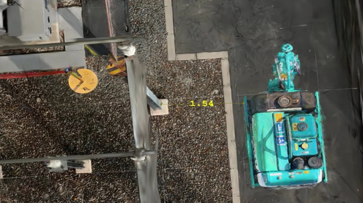 建設現場の空撮画像で、緑色の重機が黒い舗装路に停車しています。砂利の地面に「1.54」という測定値が表示されており、隣接する構造物との距離を示しているようです。