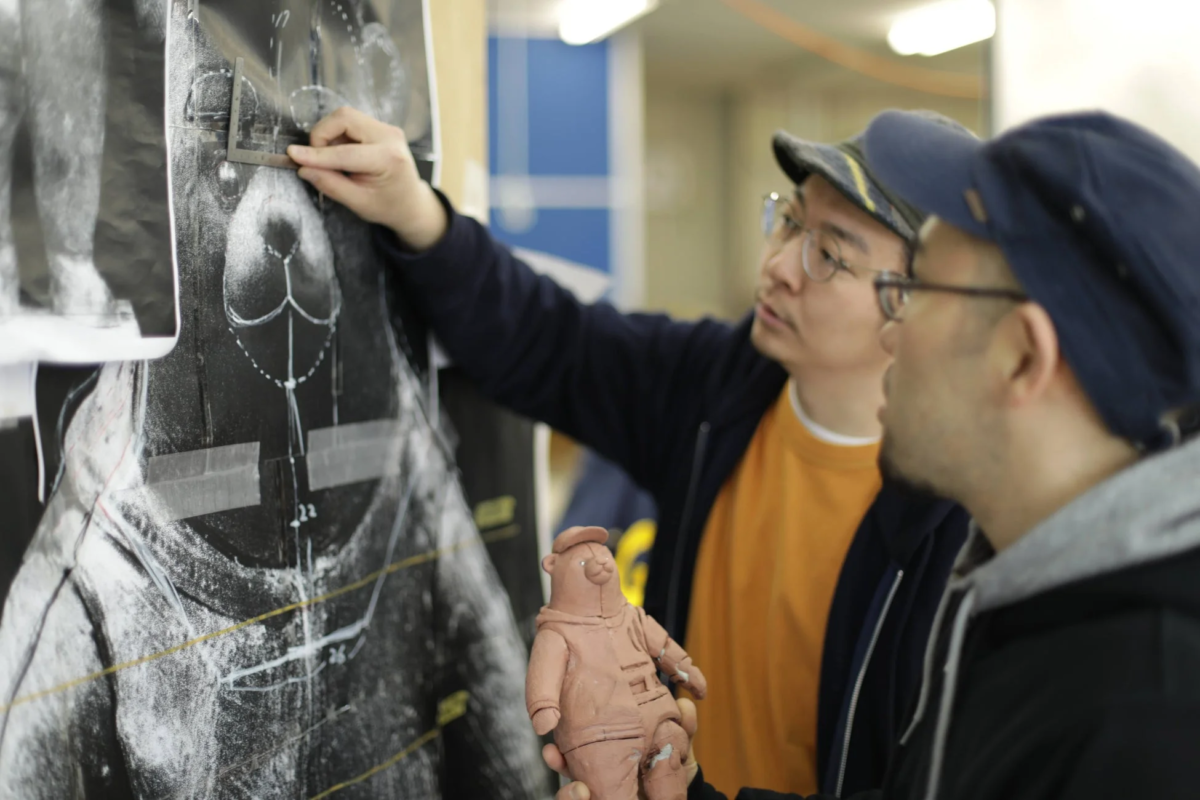 2人の男性がテディベアのようなキャラクターの大きなデザイン図面を検討している。一人は図面を指し示し、もう一人は同じキャラクターの粘土製フィギュアを持っており、制作過程での打ち合わせの様子がうかがえる。