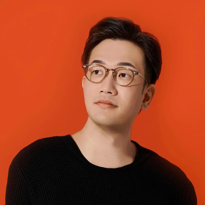 オレンジ色の背景を背に、黒いセーターと丸いメガネをかけたタマケン氏のポートレート