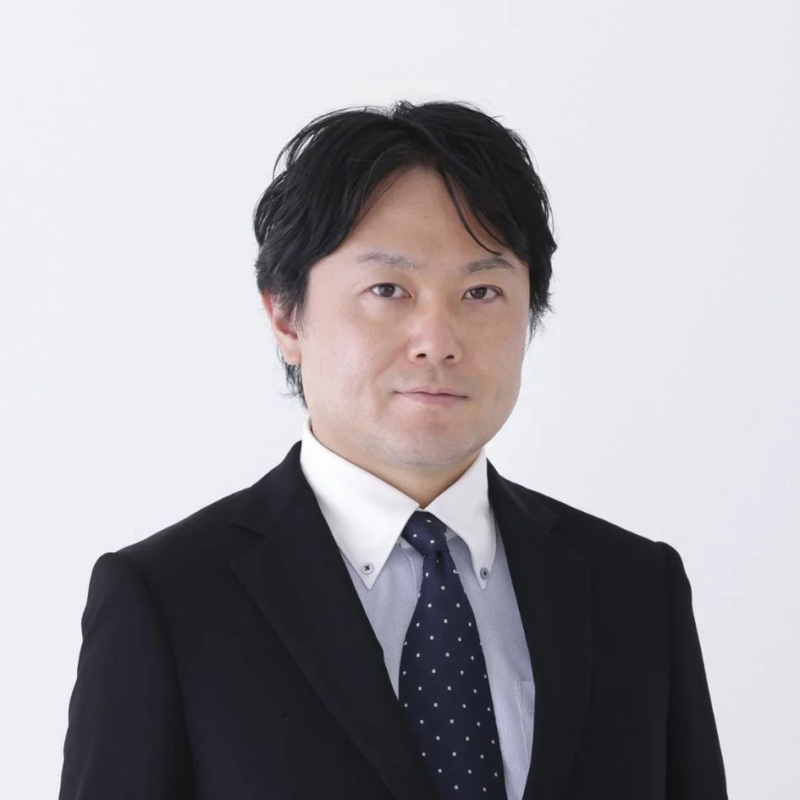 黒いスーツと水玉模様のネクタイを着用したアジア系の男性が、白い背景の前でカメラをまっすぐ見ている上半身のポートレート写真です。