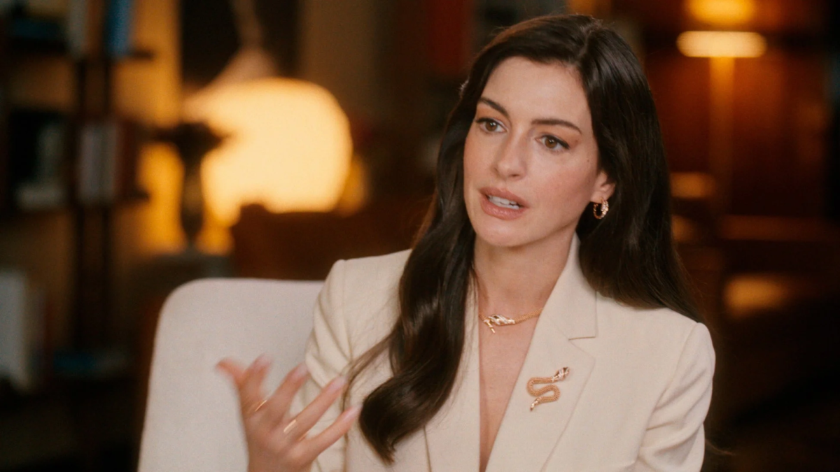 アン・ハサウェイがライトベージュのブレザーを着用し、蛇のモチーフのネックレスとブローチを身につけているポートレート。インタビュー中のような表情で、背景は暖色系の光でぼかされている。