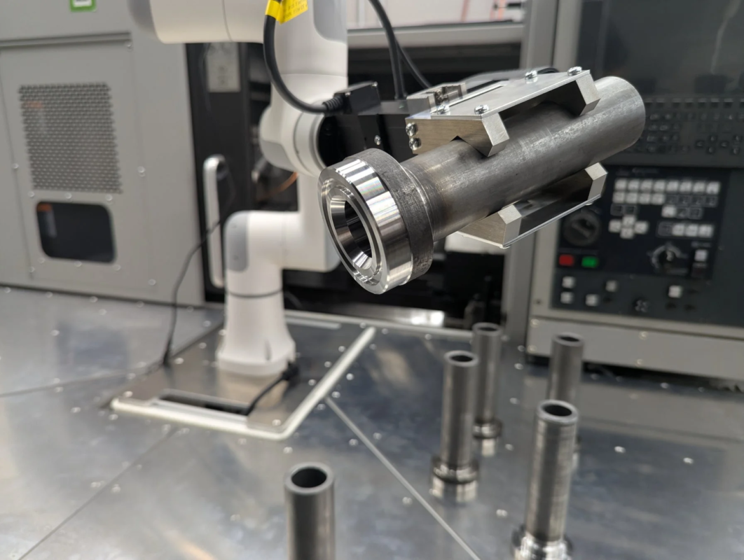 ロボットアームが金属製の円筒形部品を把持しており、自動化された製造工程の一部を示しています