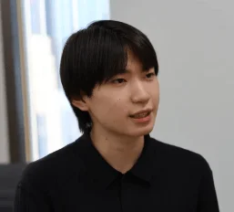 黒いシャツを着た若い男性が、室内でやや右方向を見て話しているかのような表情をしているポートレート写真です。清潔感のある短い黒髪が特徴的です。