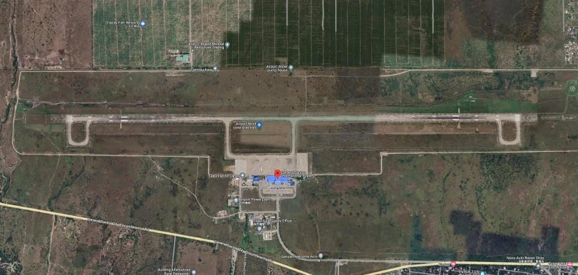 この画像は、空港とその周辺地域を示す衛星写真です。長い滑走路と複数の空港関連施設（給水所、発電所、オフィスなど）が確認でき、周囲には農地や道路、小規模な建物も点在しています。