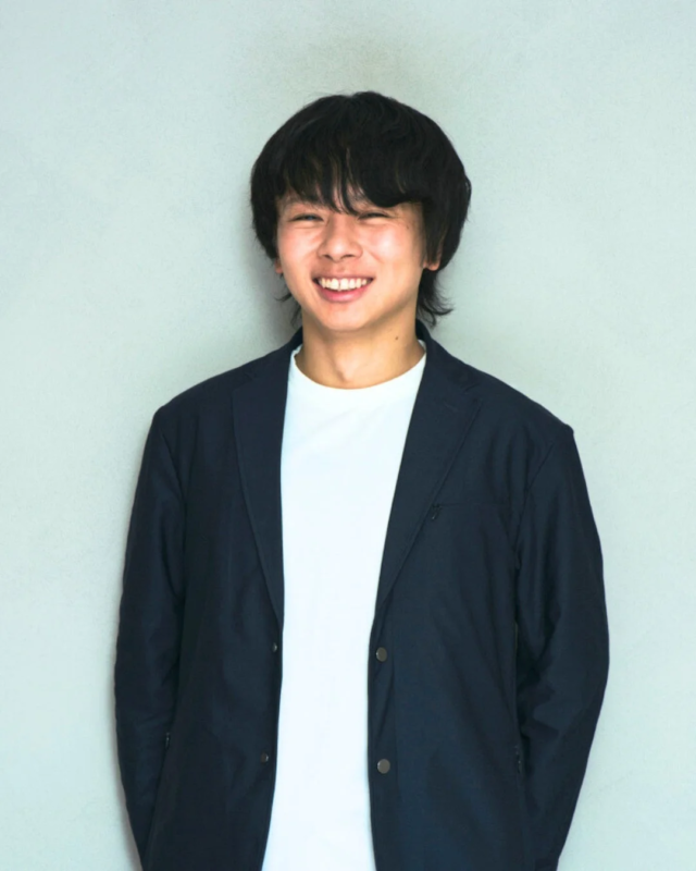 白いTシャツの上にネイビーのジャケットを羽織った若い男性のポートレート。彼は満面の笑みを浮かべ、カメラに向かって親しみやすい表情を見せています。