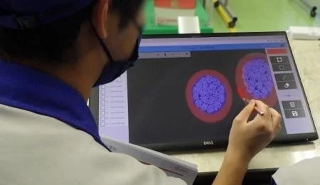 マスクと帽子を着用した人物が、タッチスクリーンモニターで細胞のような画像をスタイラスペンを使って操作しています。科学的な分析や研究の場面を示唆しています。