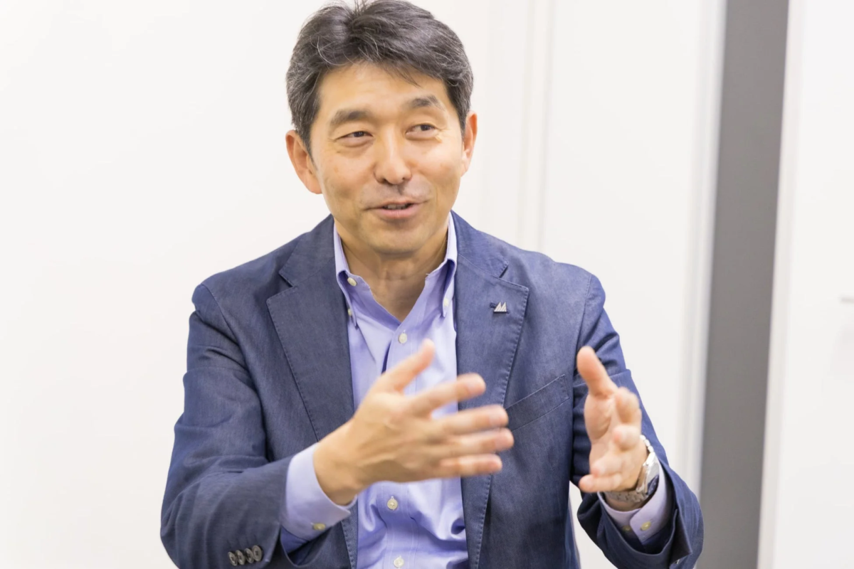 青いジャケットを着た男性が、明るい背景の前で笑顔で話しながら両手を使いジェスチャーをしています。ビジネスシーンやインタビューの一コマのようです。