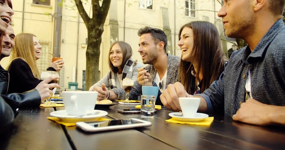 屋外のカフェテラスで、若い友人たちが集まり、笑顔で談笑しながら飲み物を楽しんでいる様子です。リラックスした雰囲気で、和やかなひとときを過ごしています。