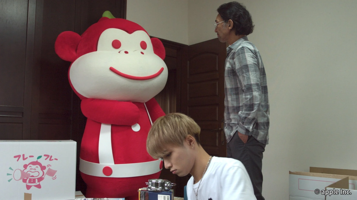 赤い猿の大きな着ぐるみと2人の男性が室内にいる。若い男性は手元の作業に集中し、年配の男性は立ち尽くしている。手前には段ボール箱も見られる。