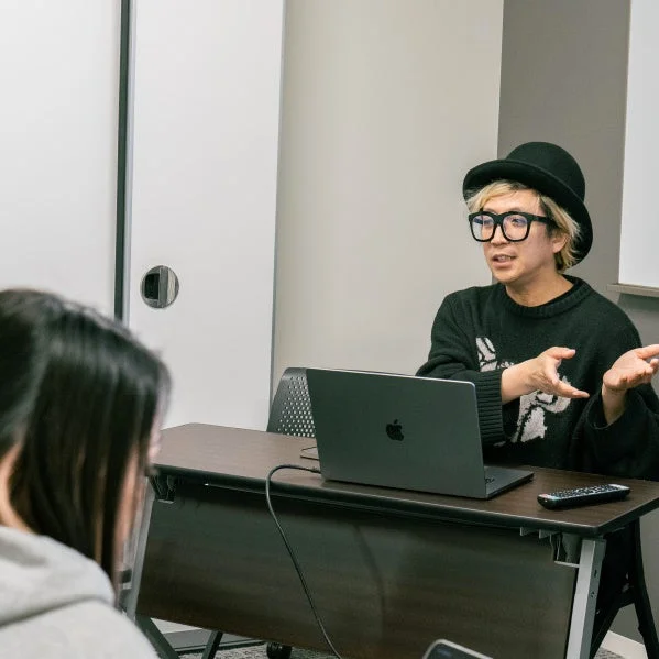 黒い帽子とメガネをかけた人物が、ノートパソコンのある机の前で聴衆に向かって話している様子