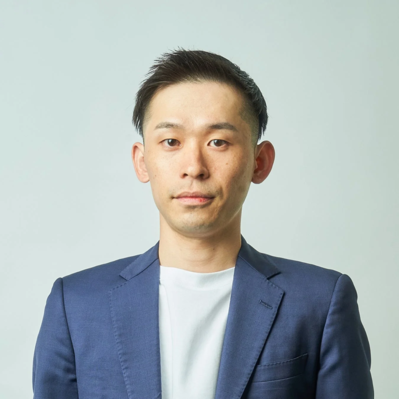 白い背景の前に立つ、ネイビーのジャケットと白いTシャツを着用したアジア系の若い男性の胸から上のポートレートです。彼はカメラを真っ直ぐに見つめています。