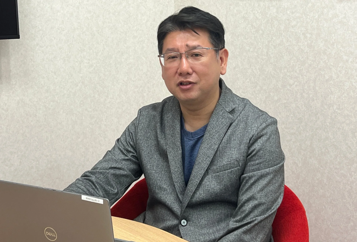 グレーのジャケットを着て眼鏡をかけた男性が、ノートパソコンの前で座っています。会議中かオフィスでの作業中の様子です。