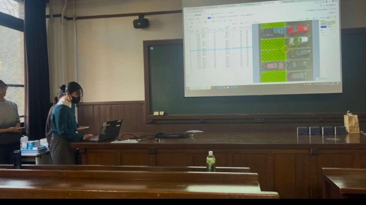 教室で女性がノートPCを操作し、プロジェクターで駐車場データと車両画像を映し出しプレゼンテーションをしている。画面にはデータ表と、緑色でハイライトされた駐車車両が表示されており、データ分析やシステムの説明をしている様子。