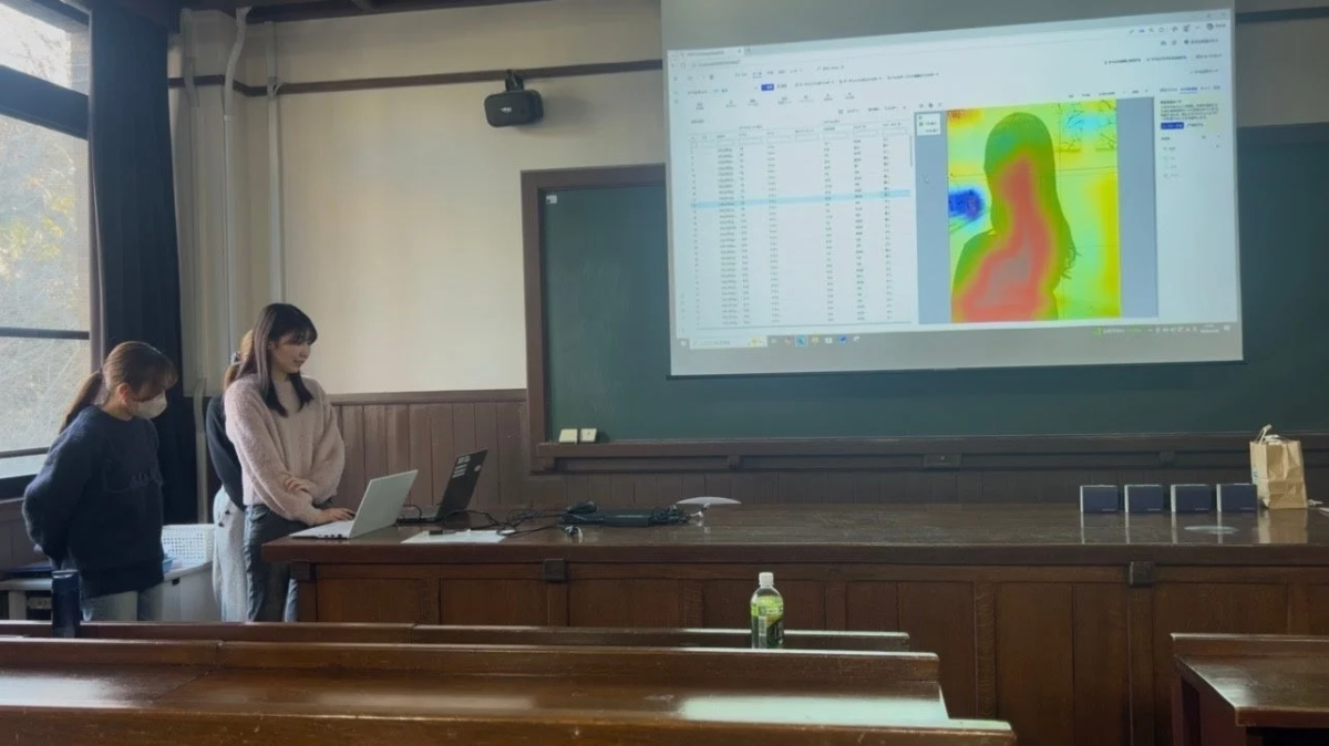 大学の教室で、2人の女性がノートパソコンを使い、プロジェクターには数値データと人物のヒートマップが投影されています。学術的な発表や研究風景のようです。