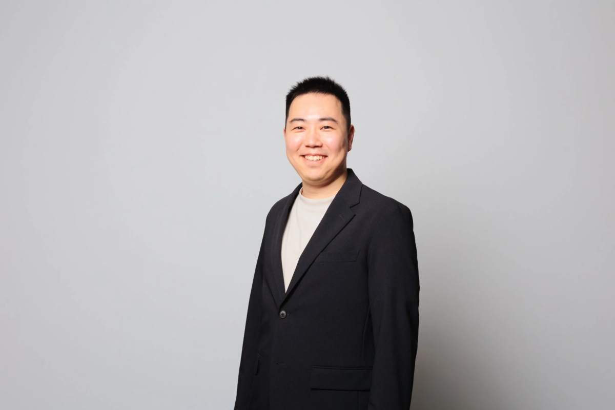 明いグレーの背景の前で、ダークスーツのジャケットとTシャツを着用したアジア系の男性が笑顔で立っているポートレート写真です。