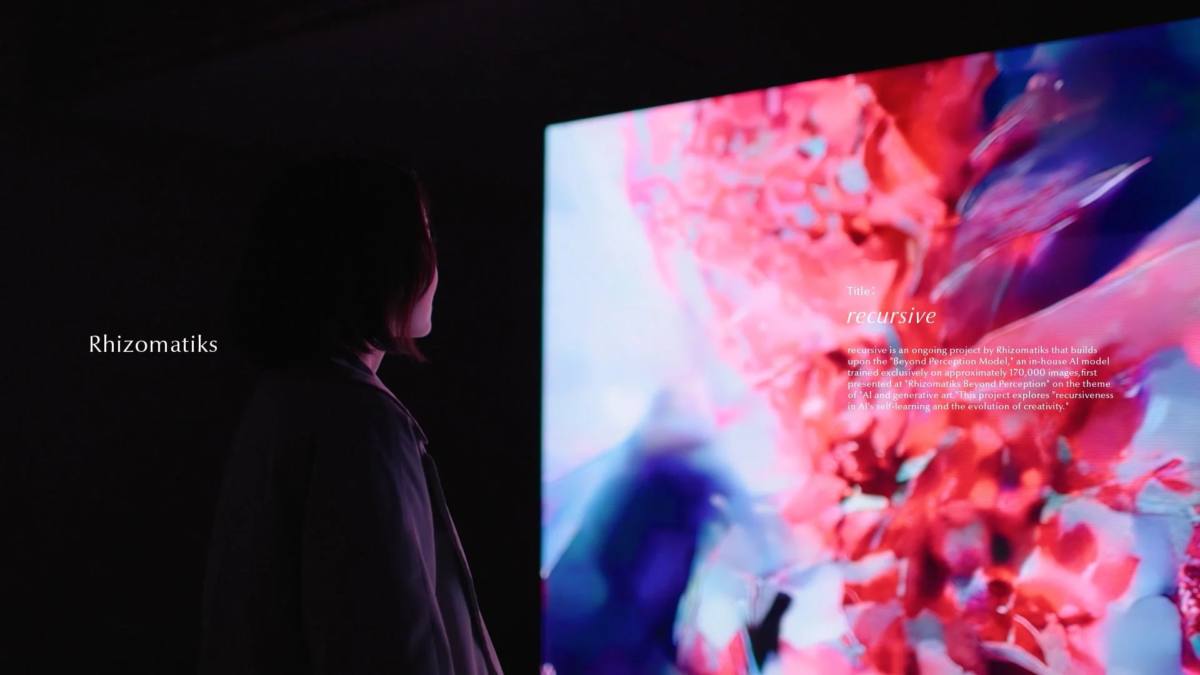 暗い空間で、人物がRhizomatiksによるAI生成アートプロジェクト「recursive」が映し出された大規模スクリーンを鑑賞している。ピンクと青の抽象的なデジタルアートが特徴的。