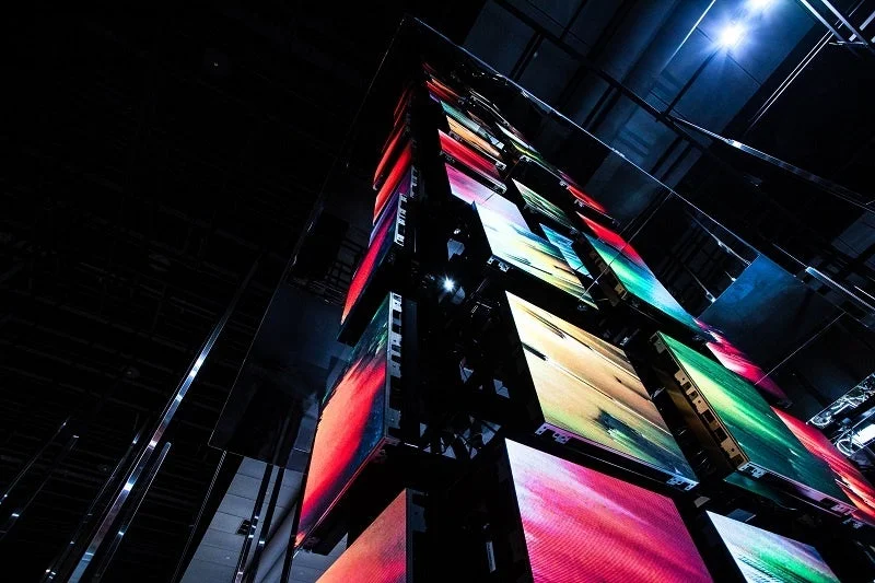 暗い空間にそびえ立つ、複数のLEDスクリーンで構成された現代的な構造物です。各スクリーンには、赤、緑、青などの鮮やかな色彩が抽象的に描かれ、デジタルアートインスタレーションのような幻想的な雰囲気を醸し出しています。