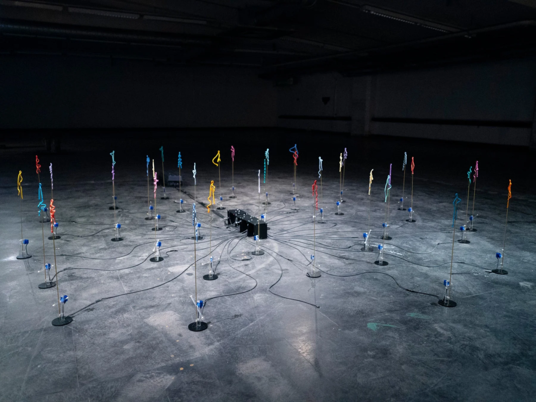 暗い広々とした空間に、多数のカラフルな人型ワイヤーアートが円形に配置され、中心の黒い装置にワイヤーで接続されている現代アートのインスタレーションです。