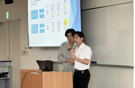 教室で2人の男性がプレゼンテーションを行っています。一人がマイクを持って話し、もう一人が隣に立っています。背後のスクリーンには図表が映し出されています。