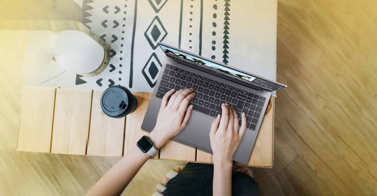 木製のテーブルでノートパソコンを操作する人物の俯瞰画像です。コーヒーカップとスマートウォッチが写り、カジュアルな作業風景を表しています。