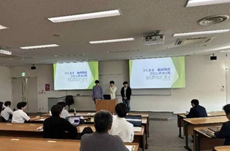 教室で学生たちが「つくるま 最終発表」と題したプレゼンテーションを行っており、他の学生が聴講している様子です。